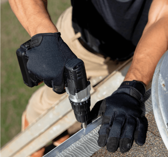 A person wearing black gloves uses a cordless power drill to install or repair a metal gutter on the edge of a roof, possibly fitting it with Gutterglove stainless steel micro-mesh. The image is taken from above, showing their arms and gloved hands.